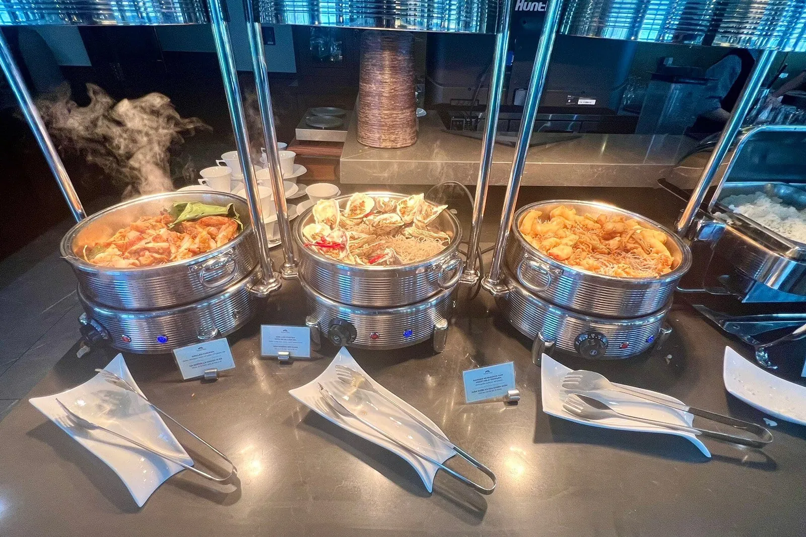 A buffet counter with three steaming chafing dishes of cooked food, serving tongs on white dishes, small name cards, a rice tray on the right, and stacked cups in the background.