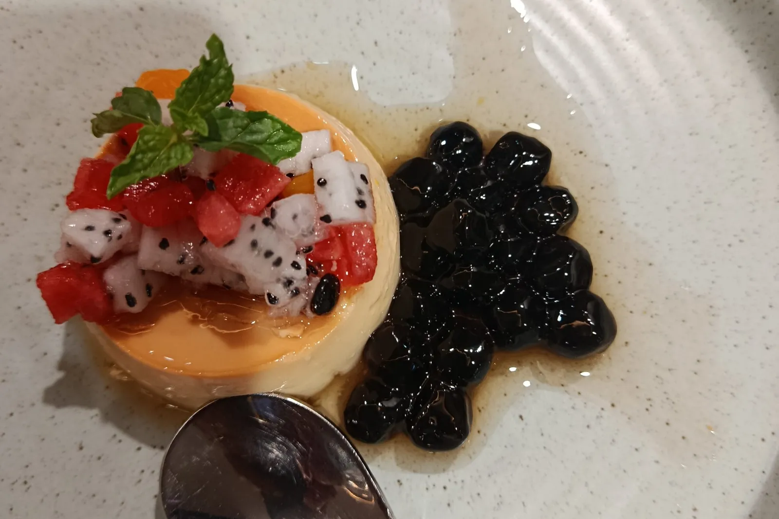 A flan-style dessert topped with dragon fruit, watermelon and mint, served with black tapioca pearls and a spoon on a speckled plate printed with 'Aspira Cruises'.