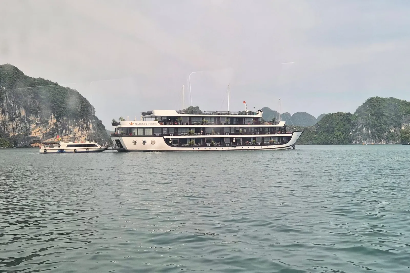 A large multi-deck white cruise ship with a small tender boat beside it sits on green-grey water in front of forested limestone karst islands under a hazy sky, photographed through what looks like a window.